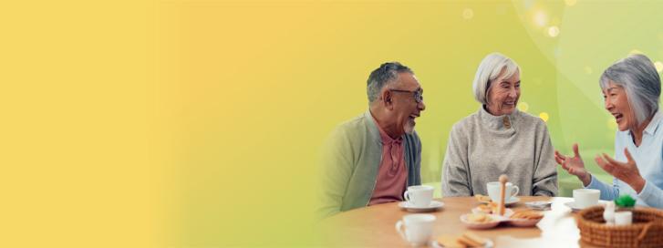 Three older adults sitting at a table, smiling and talking over tea and biscuits